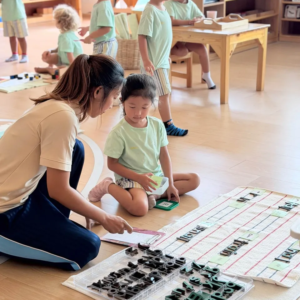 A teacher kneels beside a girl laying out wooden letters to form words on a mat