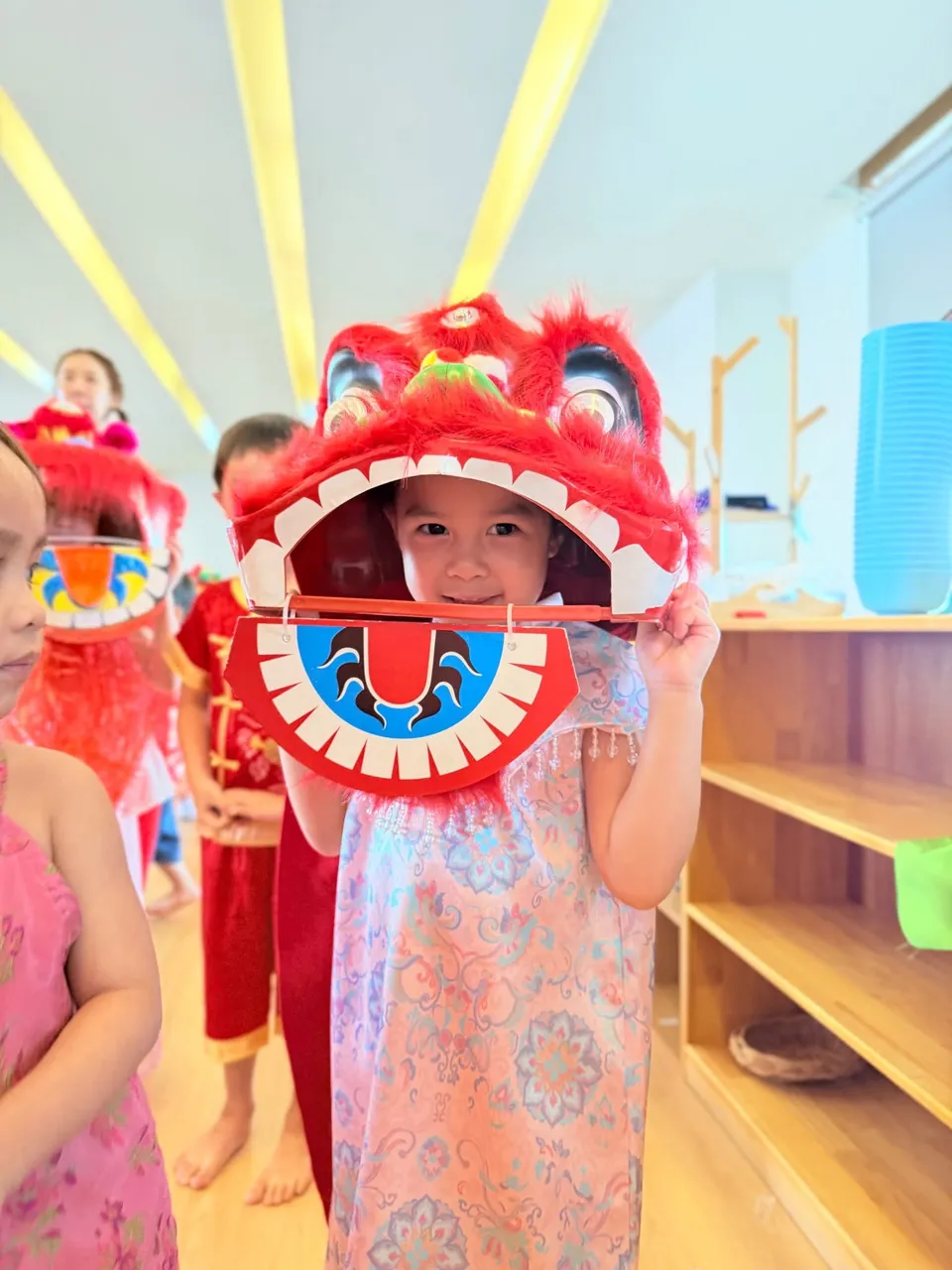 A smiling girl peeks out from under a red lion dance mask