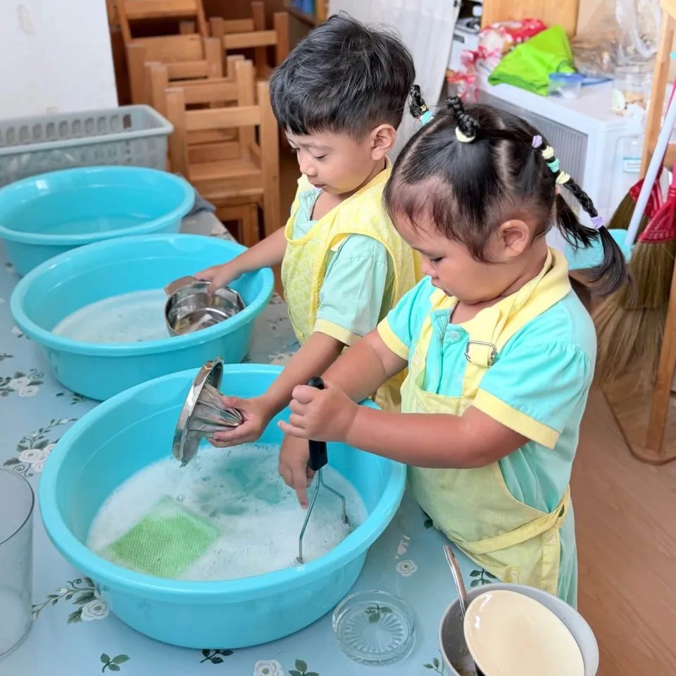 Two toddlers washing a bowl and a strainer in basins of soapy water
